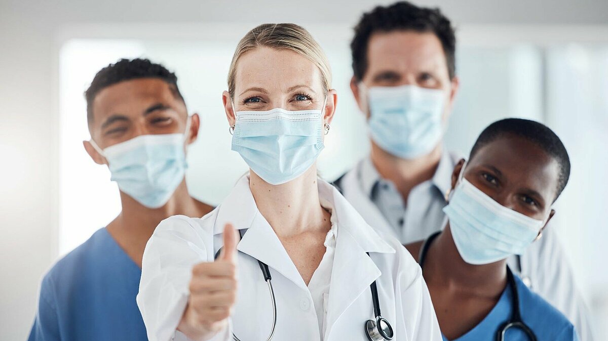 Im Bild sind eine Ärztin und vier Ärzte zu sehen. Die Ärztin steht im Vordergund vor den männlichen Kollegen und streckt den Daumen in die Höhe.